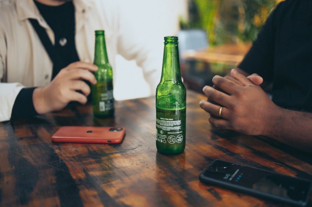 Labelled Bottles Two people enjoying drinks with green beer bottles and gadgets at a wooden table.
